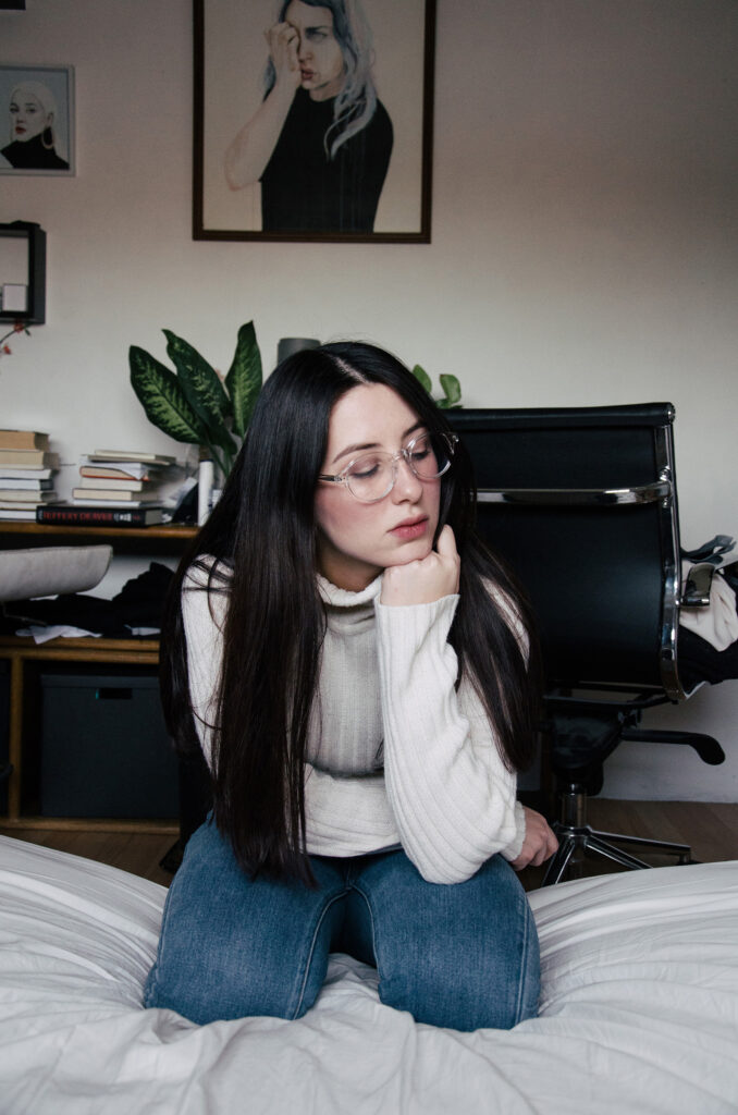 Gabriela Inzunza a thoughtful individual with glasses sits on a bed in a cozy room adorned with artwork and books.